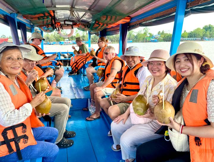 Travellers seated on a blue wooden boat enjoy fresh coconuts while cruising a broad tropical river.