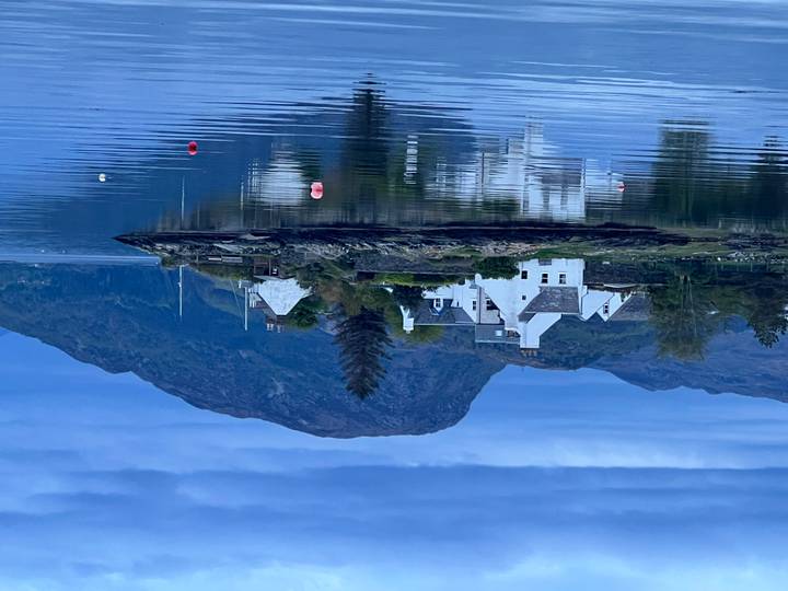 White cottages on a small peninsula reflect in the calm, dusky waters of a loch with mountains behind.