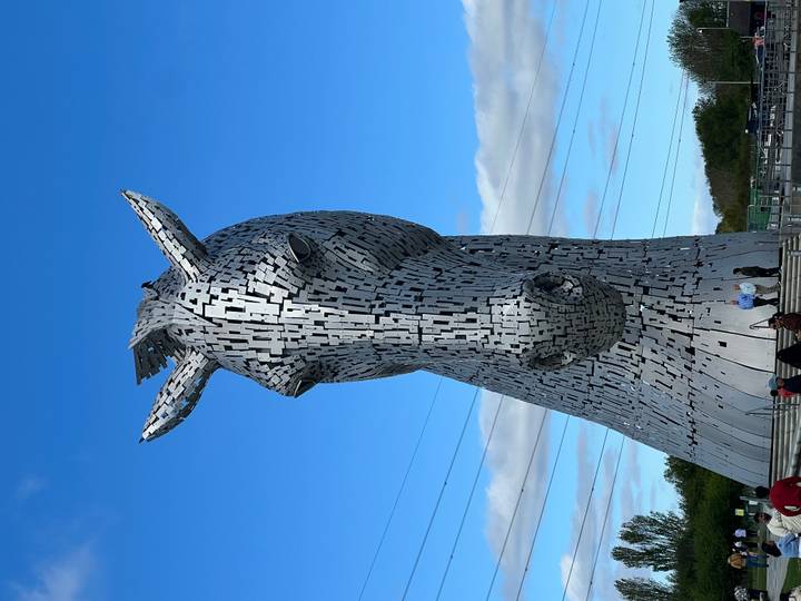 The towering metallic Kelpie horse sculpture rises against a clear blue sky with tiny visitors at its base.