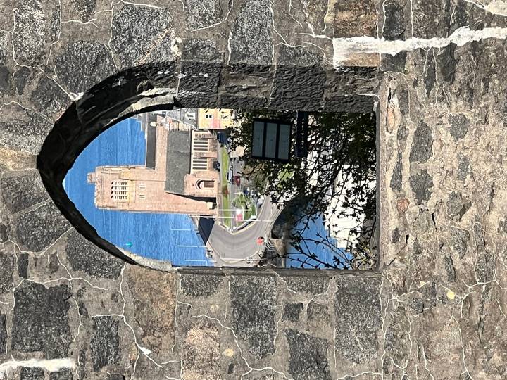 Stone window arch in a ruined wall frames a coastal church tower and winding road far below.
