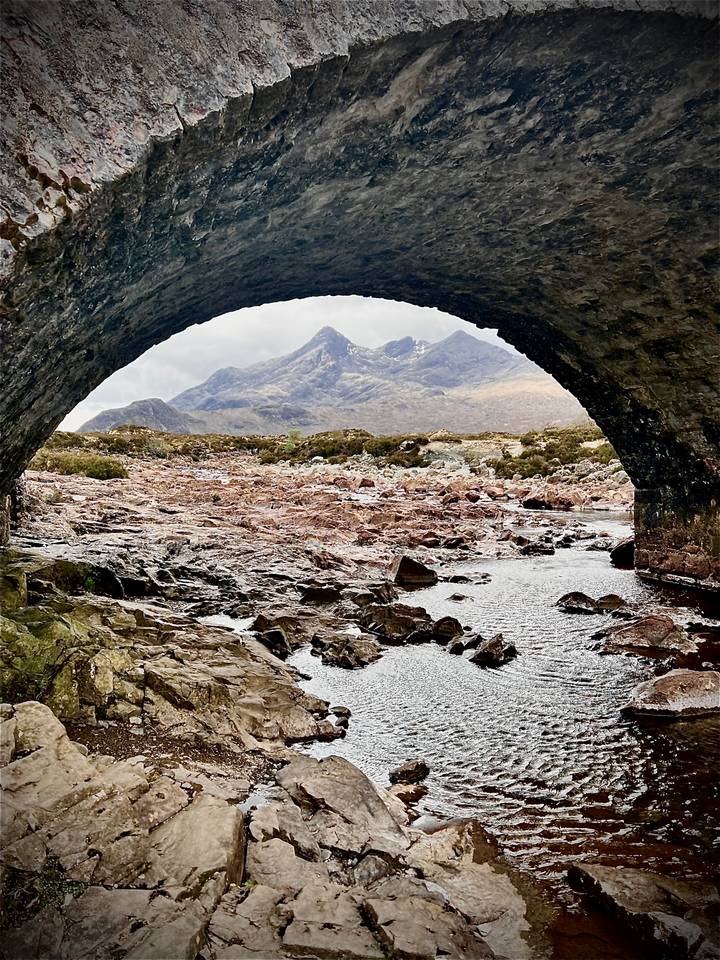 Rugged mountain peaks of the Cuillin framed beneath an old stone bridge arch with a rocky riverbed in front.