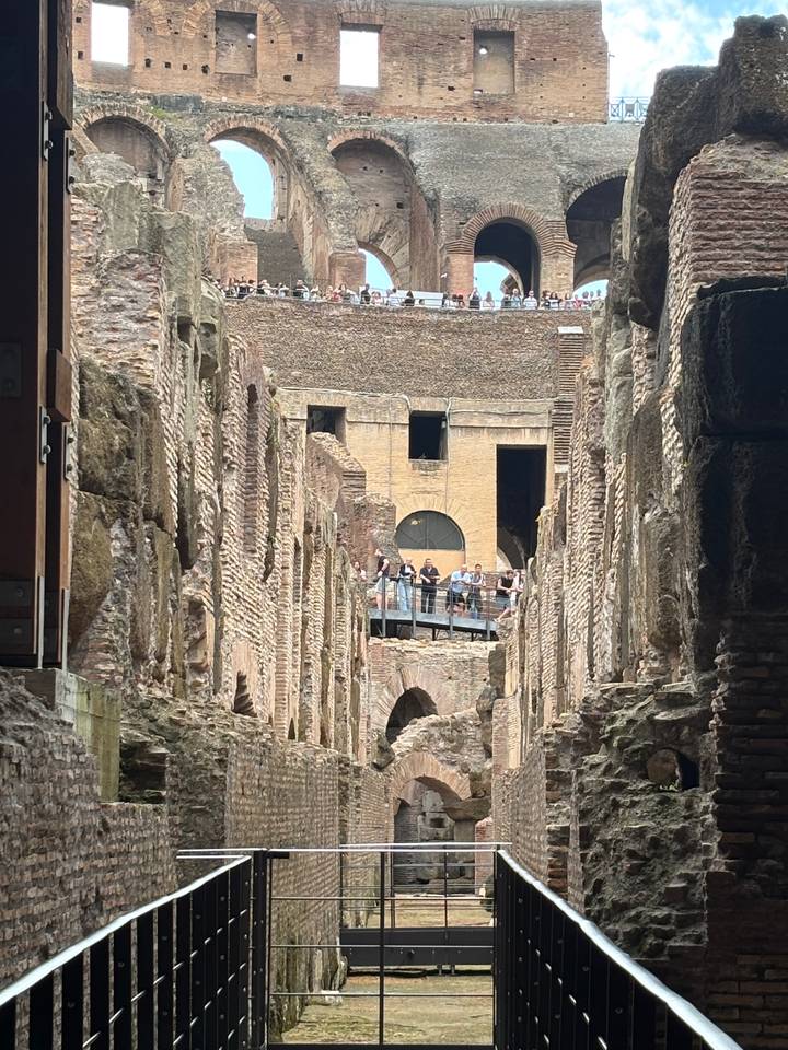 Tourists stand on a metal walkway inside ancient brick corridors of a large Roman amphitheatre.