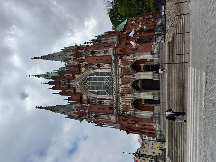 Tall red-brick gothic church with spired towers rising against a cloudy sky, pedestrians at the steps.