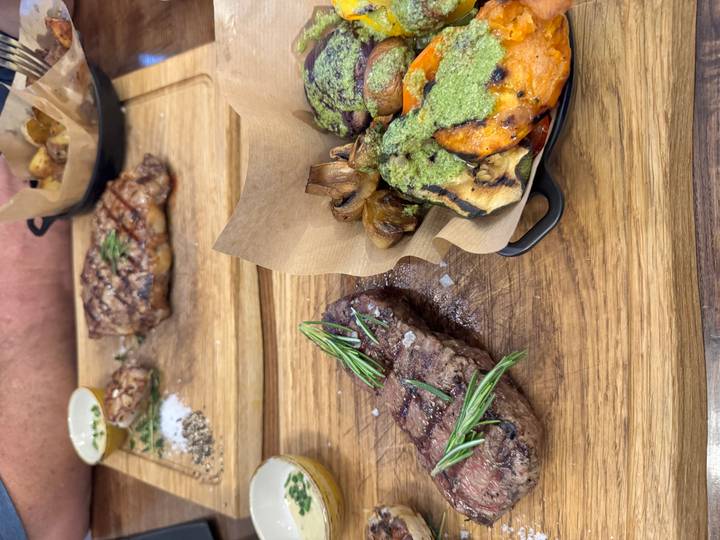 Grilled steak garnished with rosemary alongside roasted vegetables coated in green sauce on wooden boards.