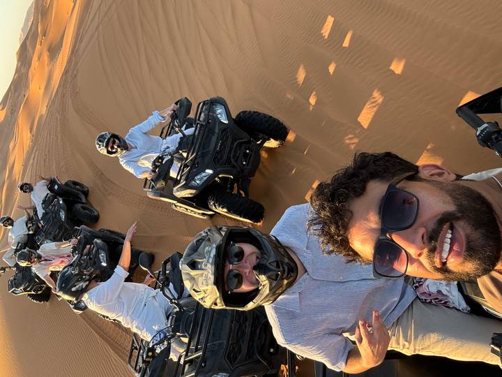 Group selfie while riding quad bikes in golden desert dunes during late afternoon light.