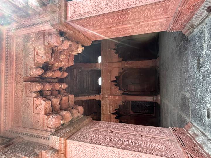 Red sandstone hall with ornate brackets and a shaft of sunlight inside a Mughal-era palace.