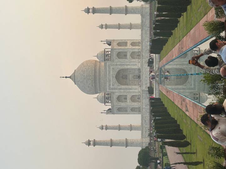 Iconic Taj Mahal framed by gardens and reflection pool at hazy sunrise with many tourists.