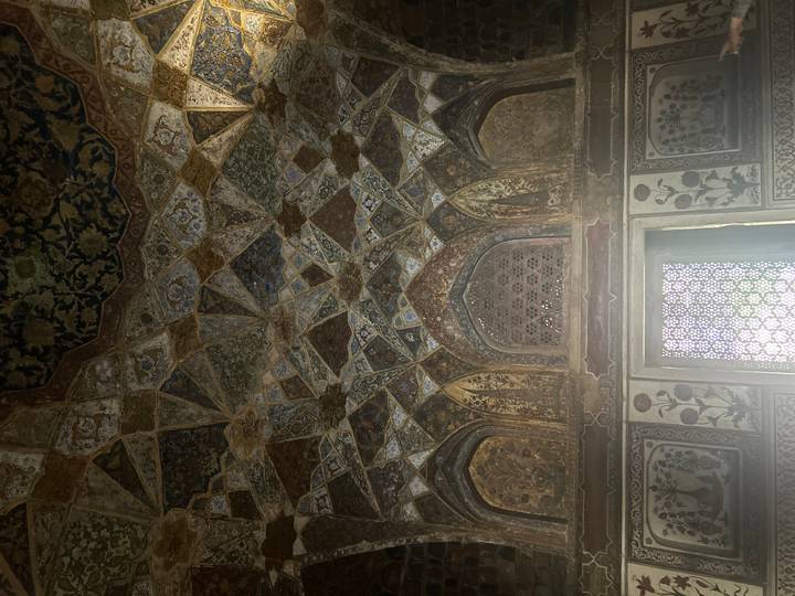Intricate geometric and floral ceiling inside a historic Mughal palace, dimly lit by window light.