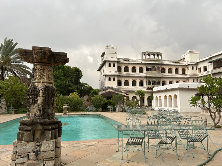 Heritage palace hotel with pool and metal garden furniture under an overcast monsoon sky.