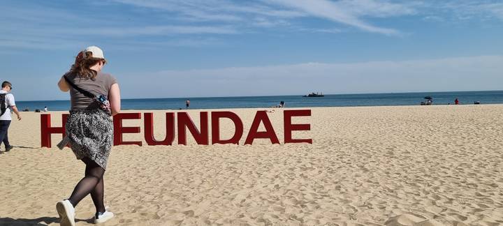 Tourist walking past red Haeundae beach sign on wide sandy shore with calm sea
