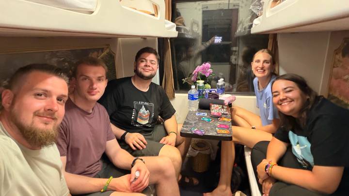 Friends relax in a cosy sleeper-train cabin, smiling around a small table with snacks and cards.