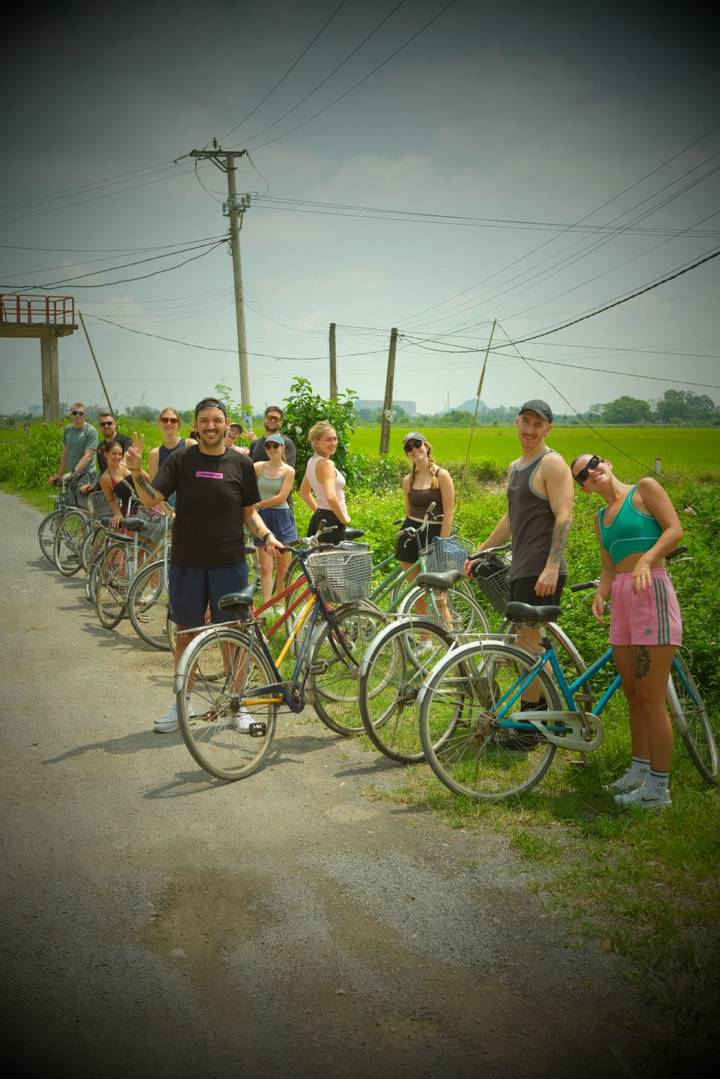 Cyclists line up with their bikes on a rural road bordered by green rice fields.
