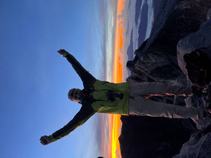 Triumphant climber raising arms on a summit at colourful dawn with valleys blanketed by clouds.