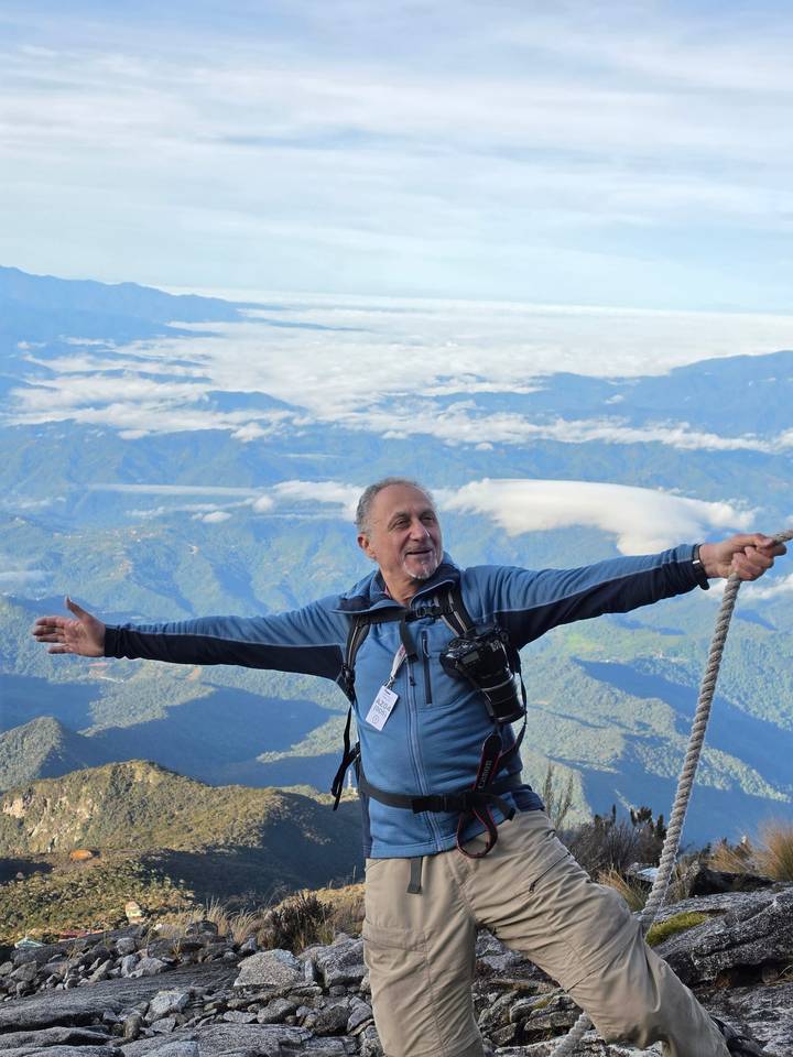 Elated climber spreads arms wide on a rugged summit ridge with colourful sunrise sky behind.