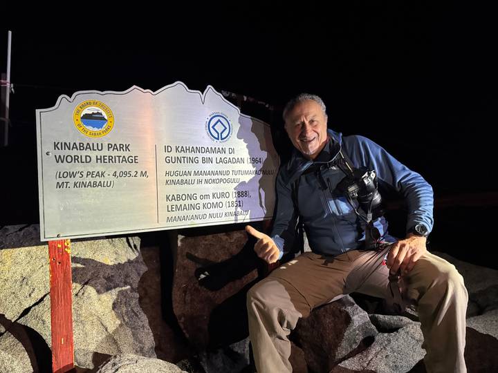 Traveller giving thumbs-up beside Kinabalu Park World Heritage stone signboard at night.