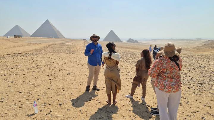 Guide explaining history to a small tour group on the desert plain with Giza pyramids in the distance.