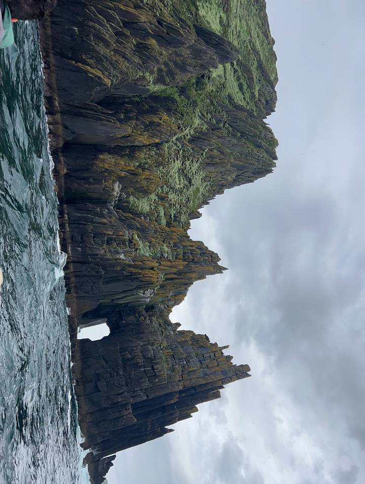 Steep, moss-covered sea stacks with a natural arch rise from choppy waters beneath a grey sky