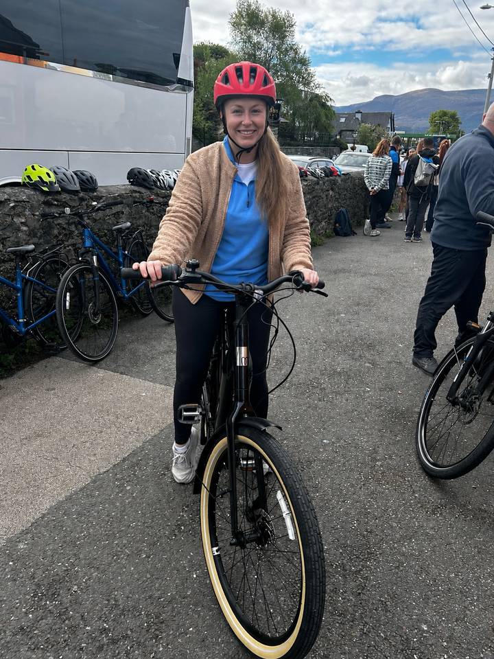 Young woman smiles while holding an e-bike in a paved courtyard surrounded by other bicycles