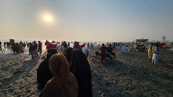 Crowds gather on a broad beach at sunset with a hazy sun low over the horizon and long shadows on wet sand.