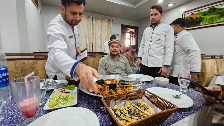 Restaurant staff in white jackets serve a large mixed grill platter to a seated diner inside an eatery.