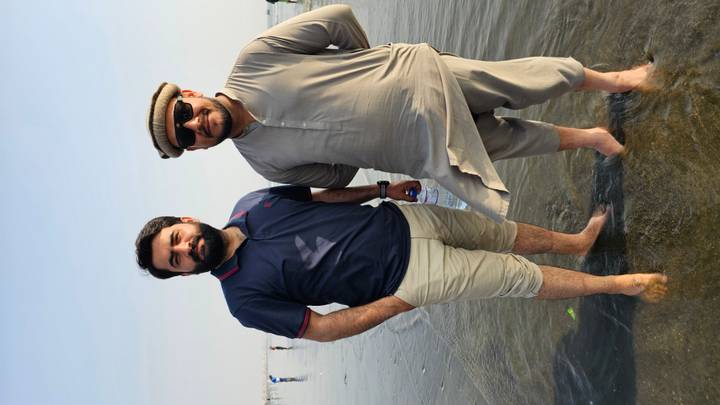 Two casually dressed men stand barefoot in shallow coastal water on a bright day.