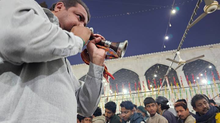 At night a man blows a traditional horn while devotees gather at a festively decorated shrine courtyard.