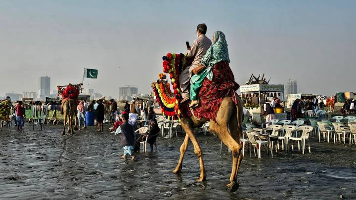 A couple in colorful attire ride a decorated camel across a busy city beach as crowds enjoy the shore.