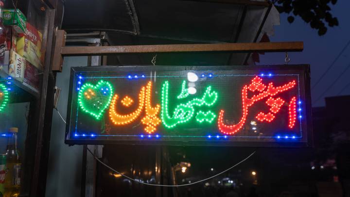 A glowing LED Urdu sign with multicolored lights hangs outside a shop after dusk.