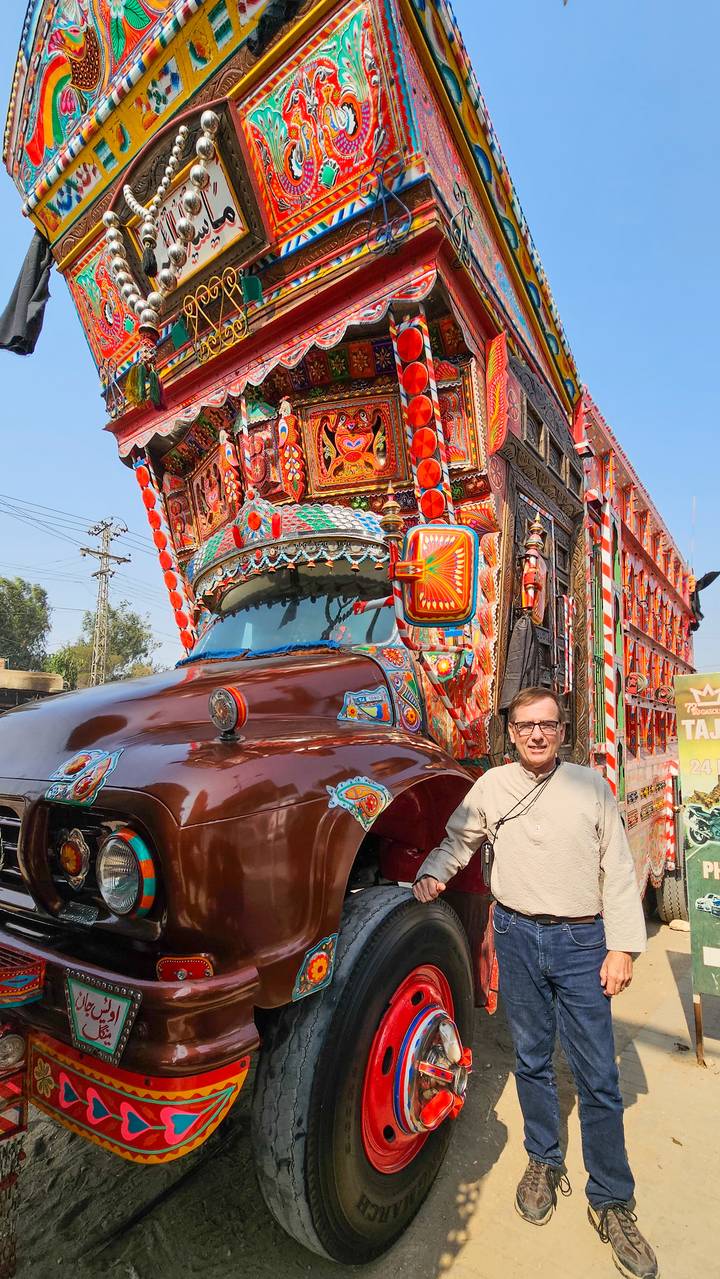 A tourist poses in front of a richly ornamented brown Pakistani cargo truck under bright sun.