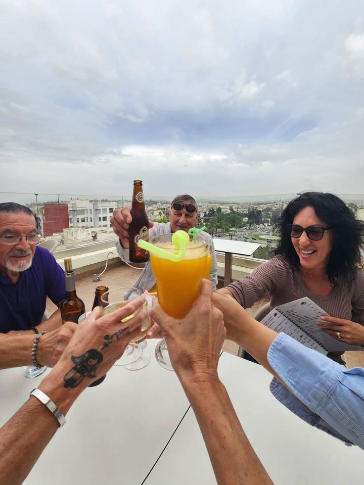 Friends raise drinks in a rooftop toast overlooking a cityscape under cloudy skies.