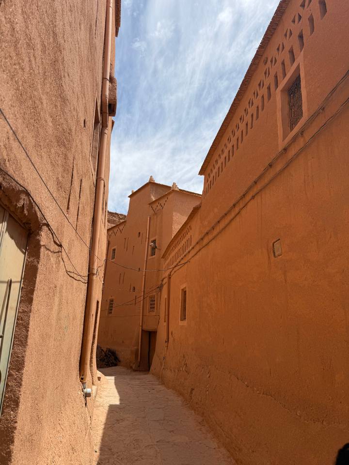 Sun-lit narrow alley lined with earthen kasbah walls and traditional adobe architecture.