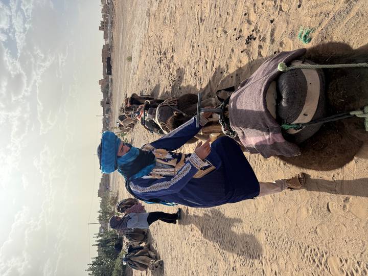 A guide in blue turban prepares camels lined up on the desert edge ready for a trek.