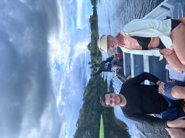 Two tourists sit in the front of a motorboat while their guide pilots along a broad jungle river beneath dramatic clouds.