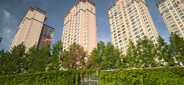 Motion-blurred photo of tall residential high-rises behind a green hedge under blue sky.