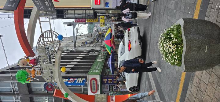 Bustling pedestrian shopping street with an arched sign, pedestrians, a white sedan and colourful umbrellas among mid-rise buildings
