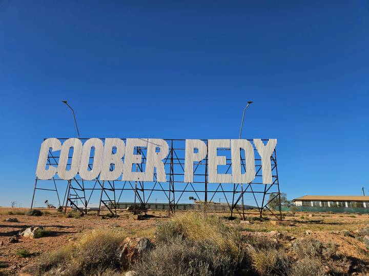 Large white metal sign spelling 'COOBER PEDY' stands against a vivid, cloudless blue sky in the outback.