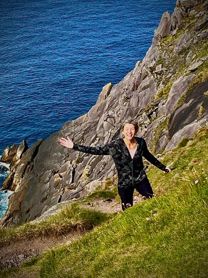 Smiling hiker poses on a green cliff edge high above the deep blue Atlantic Ocean.