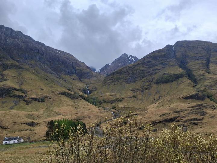 Cloudy view of a rugged Scottish mountain valley with a small waterfall trickling down rocky slopes covered in brown-green grass.