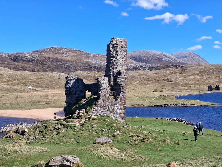 Ruined stone tower on a grassy peninsula beside a blue loch, with rolling brown hills under a clear sky.