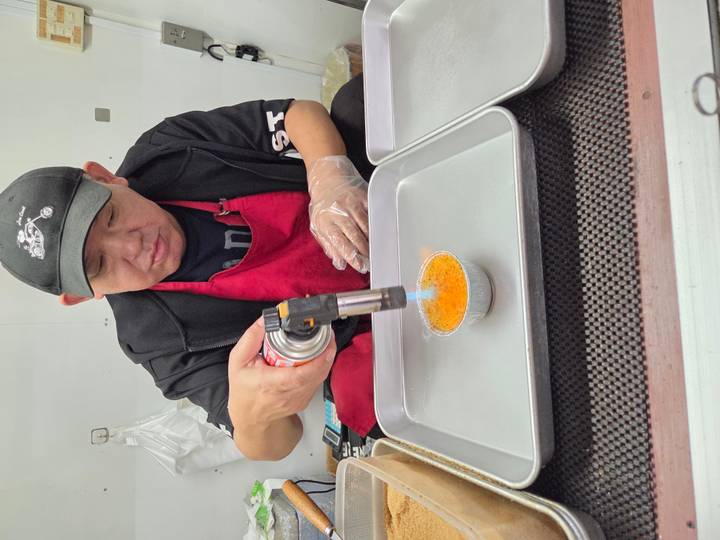 Street-food vendor in apron uses a blowtorch to caramelize a custard dessert.