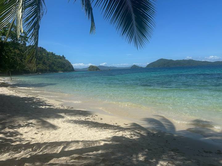 Tropical white-sand beach shaded by palm fronds with clear turquoise water and forested islets beyond.