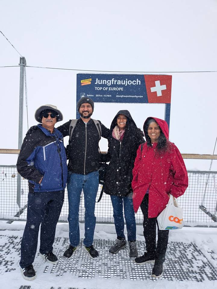 Family bundled in winter jackets smiles beside a 'Top of Europe' sign during falling snow.