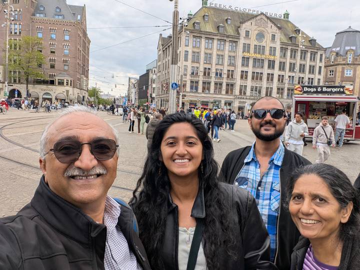 Smiling family takes a selfie in Amsterdam’s bustling Dam Square filled with trams and pedestrians.