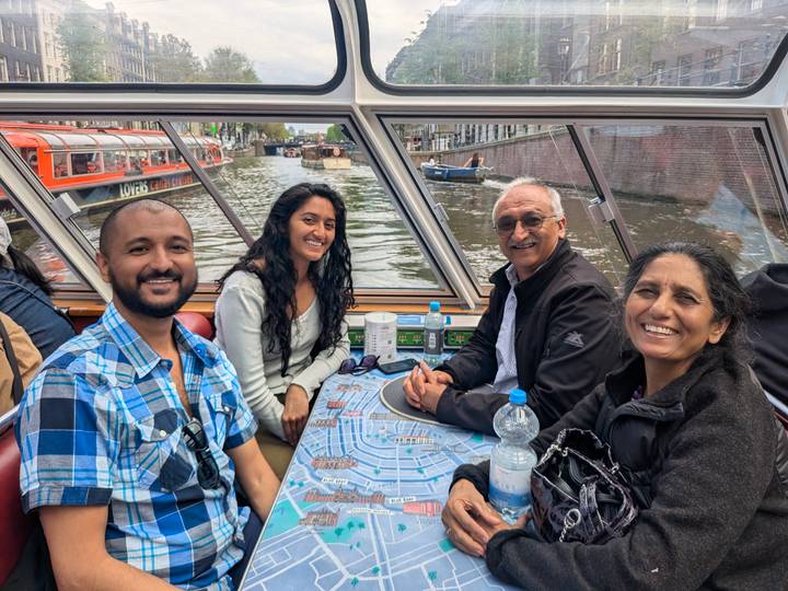 Family sits around a map-topped table inside a glass-roof canal boat cruising Amsterdam’s waterways.