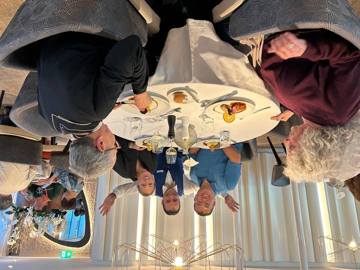 Group of travelers and a waiter cheerfully posing around a round table set with lunch dishes.