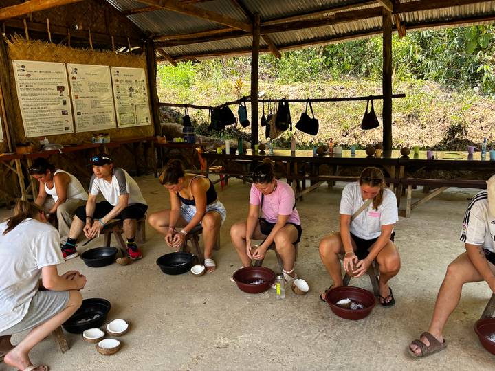 Small group of travellers seated under a thatched shelter learning to grind coconut using bowls.
