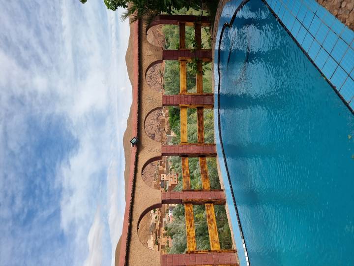 Outdoor pool framed by brick arches overlooking a river valley and rugged mountains under a partly cloudy sky.