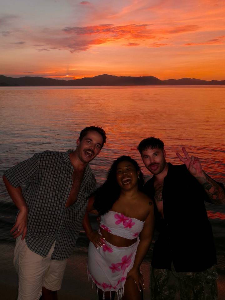 Three friends posing and laughing in front of vivid orange sunset over calm sea