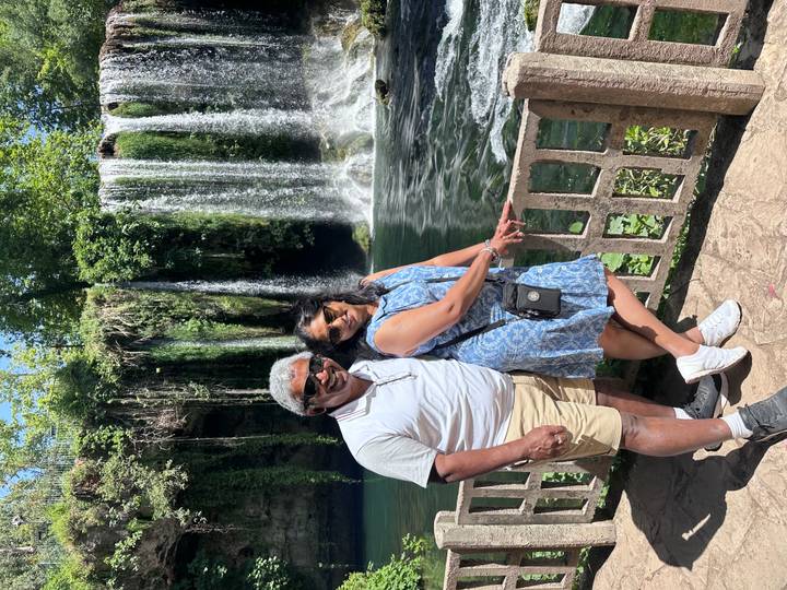 Smiling pair standing by wooden railing with lush waterfall cascading into pool behind