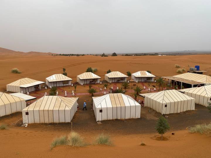 Neat rows of white canvas tents form a desert camp set amid reddish sand with distant dunes.